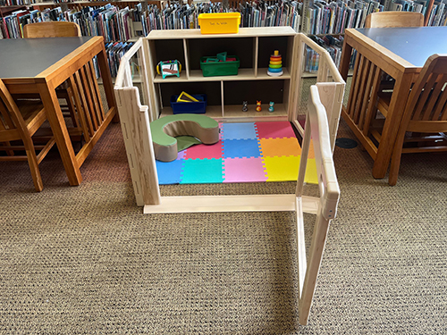 A shelf of baby toys with is the back wall of a play pen with clear sides.